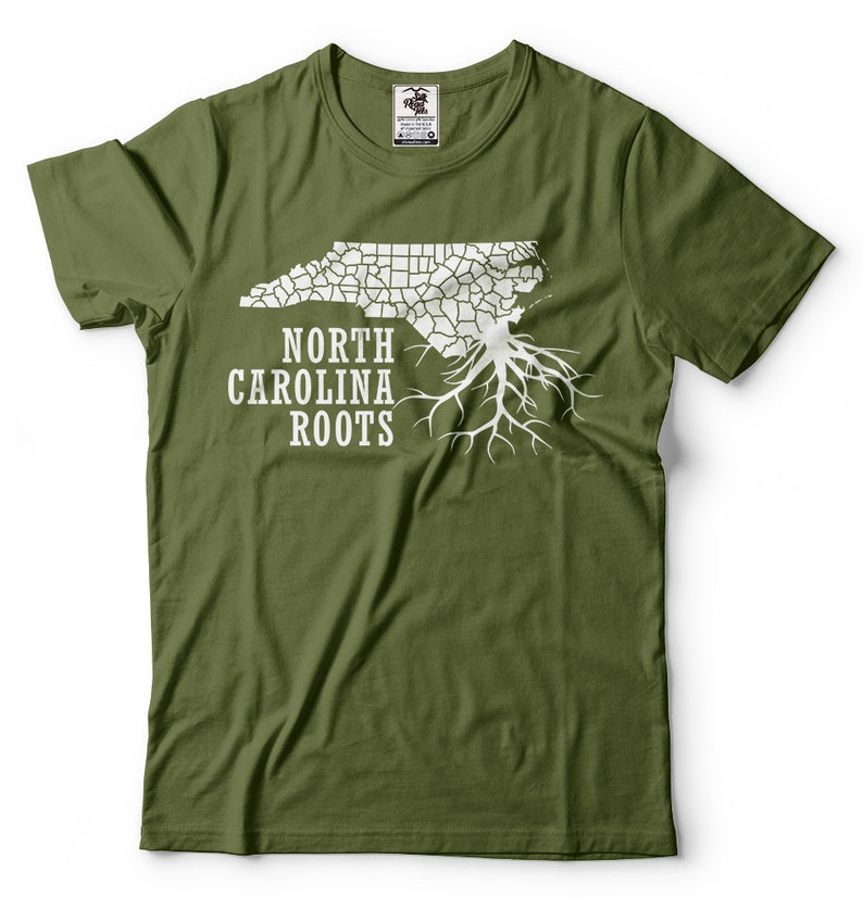 Puede incluir: Una camiseta verde con un contorno blanco del estado de Carolina del Norte y un dise&ntilde;o de ra&iacute;z de &aacute;rbol. El texto "North Carolina Roots" est&aacute; impreso debajo del contorno del estado.