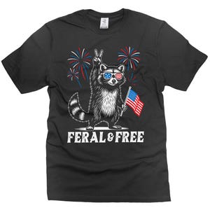 Puede incluir: Camiseta negra con un mapache con gafas de sol con el diseño de la bandera estadounidense, sosteniendo una pequeña bandera estadounidense y haciendo el signo de la paz. Fuegos artificiales en el fondo. El texto "FERAL & FREE" está debajo.