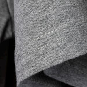 May include: Close-up of gray fabric with visible stitching. The fabric appears to be a soft knit material, with a slightly textured surface. The stitching is white and runs horizontally across the fabric.