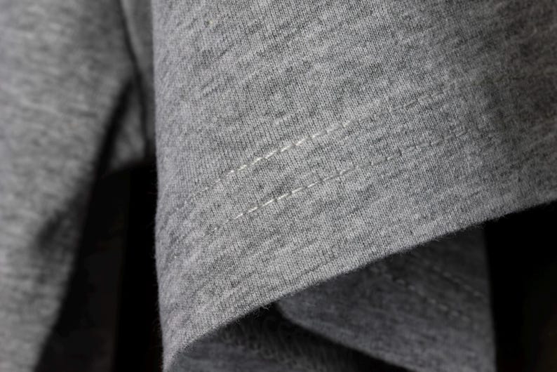 May include: Close-up of gray fabric with visible stitching. The fabric appears to be a soft knit material, with a slightly textured surface. The stitching is white and runs horizontally across the fabric.