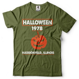K&ouml;nnte beinhalten: Gr&uuml;nes T-Shirt mit einer wei&szlig;en Grafik, auf der "HALLOWEEN 1978 HADDONFIELD, ILLINOIS" steht, mit einem roten und orangefarbenen K&uuml;rbis in der Mitte.