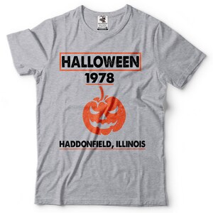 K&ouml;nnte beinhalten: Graues T-Shirt mit einem schwarz-wei&szlig;en Grafikdruck, auf dem "HALLOWEEN 1978 HADDONFIELD, ILLINOIS" steht. Die Grafik enth&auml;lt auch einen orangefarbenen K&uuml;rbis mit einem gruseligen Gesicht.