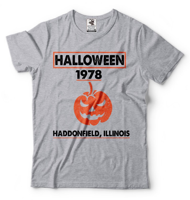 K&ouml;nnte beinhalten: Graues T-Shirt mit einem schwarz-wei&szlig;en Grafikdruck, auf dem "HALLOWEEN 1978 HADDONFIELD, ILLINOIS" steht. Die Grafik enth&auml;lt auch einen orangefarbenen K&uuml;rbis mit einem gruseligen Gesicht.