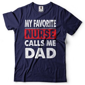 May include: Navy blue t-shirt with white text that reads "My Favorite Nurse Calls Me Dad". The word "Nurse" is in red.