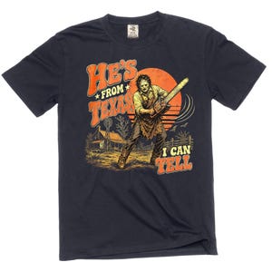 Pode incluir: Uma t-shirt de manga curta azul-marinho escuro com uma estampa gráfica de uma figura segurando uma motosserra diante de um grande sol laranja. O texto diz HE'S FROM TEXAS, I CAN TELL. A cena inclui uma casa rural, um moinho e uma cerca.
