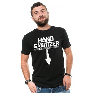 Pode incluir: T-shirt preto com as palavras "HAND SANITIZER" em letras brancas. Abaixo, uma seta para baixo e o texto "PUMP 10-15 TIMES THEN RUB IN VIGOROUSLY". A camiseta é usada por uma pessoa.