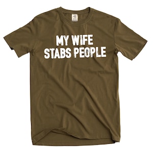 May include: An olive green t-shirt with the white text "MY WIFE STABS PEOPLE". The crew neck shirt is made of a soft material. The text is in a bold, sans-serif font and is centered on the front of the shirt.