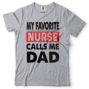 May include: Gray t-shirt with the text "My Favorite Nurse Calls Me Dad" in black and red lettering. The word "Nurse" is in a red rectangle.