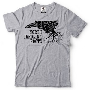Puede incluir: Una camiseta gris con un contorno negro del estado de Carolina del Norte y las palabras "North Carolina Roots" impresas en ella.