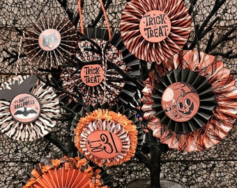 May include: A black tree branch with six Halloween-themed paper rosettes. The rosettes are orange and black with various designs, including pumpkins, bats, and the words "Trick or Treat". The tree is in a black pot and is sitting on a black and white spiderweb patterned surface.