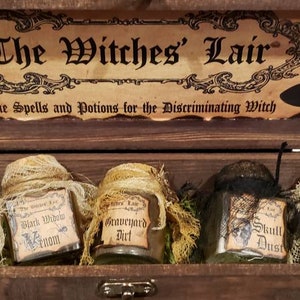 Może przedstawiać: Drewniana skrzynia z napisem "The Witches' Lair" na pokrywie. Wewnątrz skrzyni znajduje się pięć małych szklanych słoik&oacute;w z etykietami, na kt&oacute;rych widnieje napis "Eye of Newt", "Black Widow Venom", "Graveyard Dirt", "Skull Dust" i "Bat Wings".