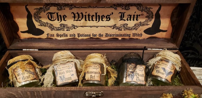 Może przedstawiać: Drewniana skrzynia z napisem "The Witches' Lair" na pokrywie. Wewnątrz skrzyni znajduje się pięć małych szklanych słoik&oacute;w z etykietami, na kt&oacute;rych widnieje napis "Eye of Newt", "Black Widow Venom", "Graveyard Dirt", "Skull Dust" i "Bat Wings".