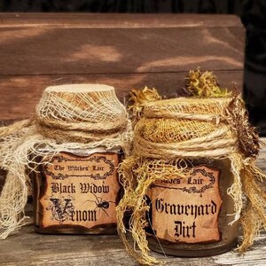 Może przedstawiać: Dwa małe szklane słoiki z korkowymi pokrywkami, owinięte w pł&oacute;tno i koronkę. Słoiki są oznaczone etykietami "The Witches Lair Black Widow Venom" i "Graves Lair Graveyard Dirt".