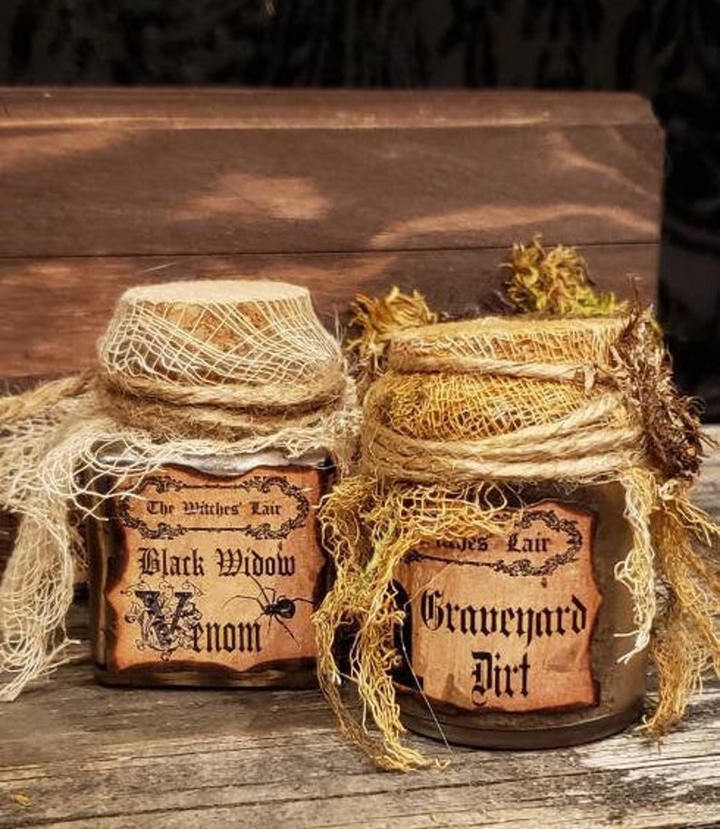 Może przedstawiać: Dwa małe szklane słoiki z korkowymi pokrywkami, owinięte w pł&oacute;tno i koronkę. Słoiki są oznaczone etykietami "The Witches Lair Black Widow Venom" i "Graves Lair Graveyard Dirt".