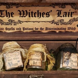Może przedstawiać: Drewniana skrzynia z etykietą z napisem "The Witches' Lair Fine Spells and Potions for the Discriminating Witch". Skrzynia zawiera sześć małych szklanych słoik&oacute;w z etykietami z napisami "Eye of Newt", "Black Widow Venom", "Graveyard Dirt", "Skull Dust", "Bat Wings" i "The Witches' Lair".