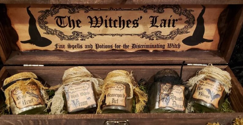 Może przedstawiać: Drewniana skrzynia z etykietą z napisem "The Witches' Lair Fine Spells and Potions for the Discriminating Witch". Skrzynia zawiera sześć małych szklanych słoik&oacute;w z etykietami z napisami "Eye of Newt", "Black Widow Venom", "Graveyard Dirt", "Skull Dust", "Bat Wings" i "The Witches' Lair".