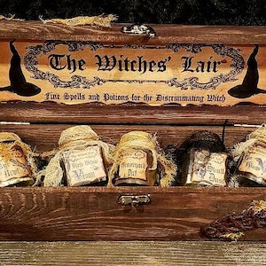 Może przedstawiać: Drewniana skrzynia z zawiasowym wiekiem, oznaczona napisem "The Witches' Lair: Fine Spells and Potions for the Discriminating Witch". Skrzynia zawiera pięć małych szklanych słoik&oacute;w z etykietami i sznurkiem owiniętym wok&oacute;ł g&oacute;rnej części.