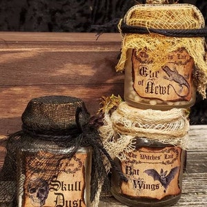 Może przedstawiać: Trzy szklane słoiki z etykietami i pokrywkami z pł&oacute;tna. Etykiety głosi: "The Witches Lair", "Skull Dust" i "Bat Wings". Słoiki zawierają białe koraliki, biały proszek i małą figurkę nietoperza.