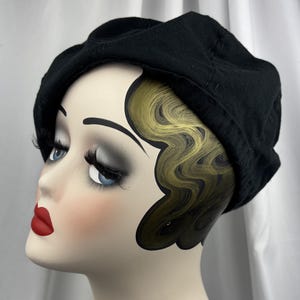 May include: A solid black beret made of a soft fabric material. The beret features a rounded crown and a structured band that sits securely around the head. The fabric has a matte finish and visible stitching details along the edge of the crown.