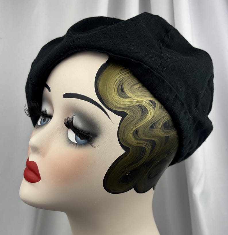 May include: A solid black beret made of a soft fabric material. The beret features a rounded crown and a structured band that sits securely around the head. The fabric has a matte finish and visible stitching details along the edge of the crown.
