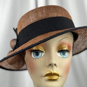 Pu&ograve; includere: Un cappello di paglia marrone con un nastro nero e un fiocco nero sul lato. Il cappello &egrave; inclinato e viene indossato da un manichino.