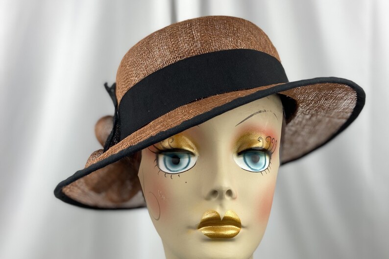 Pu&ograve; includere: Un cappello di paglia marrone con un nastro nero e un fiocco nero sul lato. Il cappello &egrave; inclinato e viene indossato da un manichino.