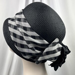 May include: A black woven hat with a wide brim and a decorative black and white checkered scarf wrapped around the crown. A black fabric flower is attached to the scarf. The hat is displayed on a mannequin head.