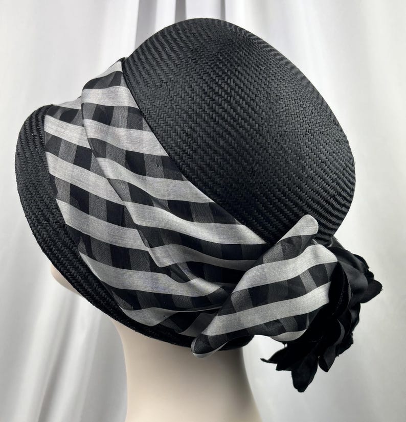 May include: A black woven hat with a wide brim and a decorative black and white checkered scarf wrapped around the crown. A black fabric flower is attached to the scarf. The hat is displayed on a mannequin head.