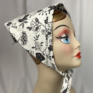 May include: A white headscarf with a black floral pattern is tied around a mannequin's head. The scarf features a repeating design of flowers and leaves. The edges of the scarf are trimmed with a black and white checkered pattern. The scarf is tied under the chin.