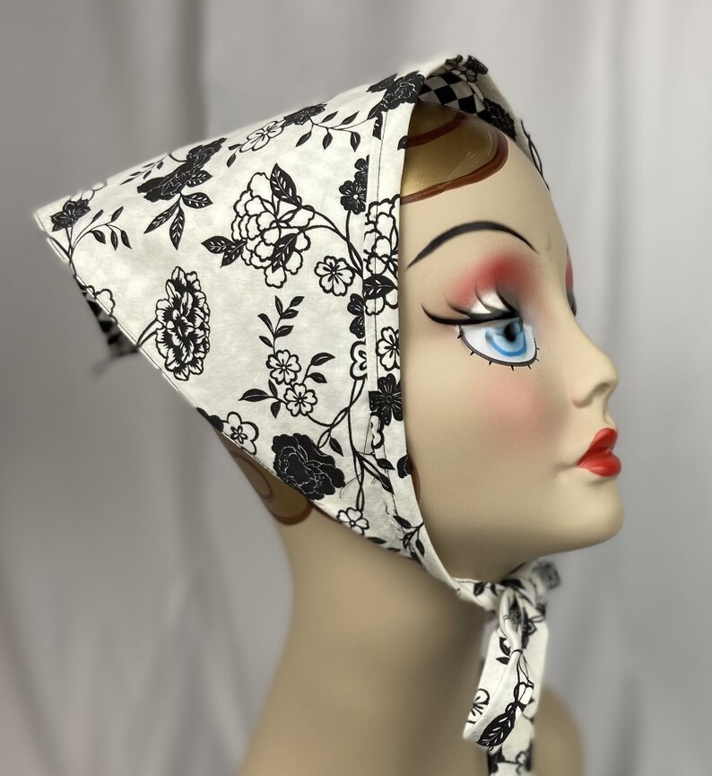 May include: A white headscarf with a black floral pattern is tied around a mannequin's head. The scarf features a repeating design of flowers and leaves. The edges of the scarf are trimmed with a black and white checkered pattern. The scarf is tied under the chin.