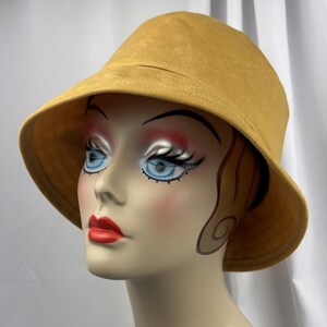 Puede incluir: Un sombrero de pescador de color amarillo mostaza con una textura suave y mate y un ala ancha inclinada hacia abajo. El sombrero presenta una copa redondeada y l&iacute;neas simples y limpias, proporcionando una cobertura total.
