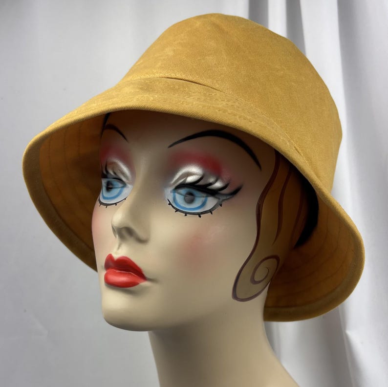 Puede incluir: Un sombrero de pescador de color amarillo mostaza con una textura suave y mate y un ala ancha inclinada hacia abajo. El sombrero presenta una copa redondeada y l&iacute;neas simples y limpias, proporcionando una cobertura total.