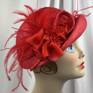 May include: A vibrant red fascinator hat featuring a woven straw texture, adorned with a large satin ribbon bow, fabric roses, and long, wispy red feathers extending from the side. The hat is displayed on a mannequin head profile.