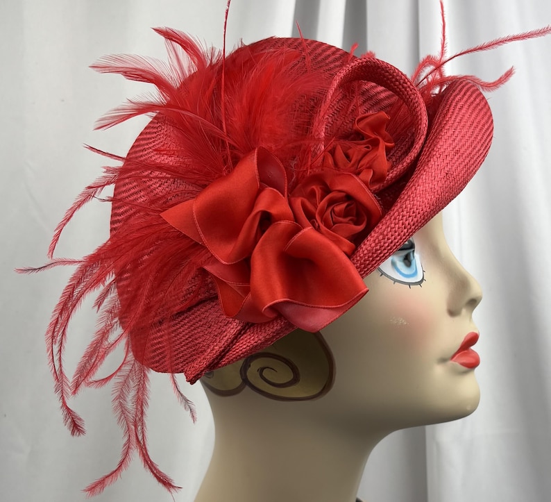 May include: A vibrant red fascinator hat featuring a woven straw texture, adorned with a large satin ribbon bow, fabric roses, and long, wispy red feathers extending from the side. The hat is displayed on a mannequin head profile.