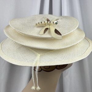 May include: A cream-colored straw hat featuring a tiered, multi-layered brim design. A decorative dragonfly ornament with brown and cream wings and a green eye is attached to the top layer. Two thin, braided cream strings hang down.