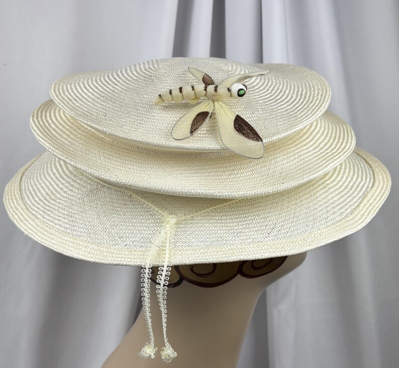 May include: A cream-colored straw hat featuring a tiered, multi-layered brim design. A decorative dragonfly ornament with brown and cream wings and a green eye is attached to the top layer. Two thin, braided cream strings hang down.