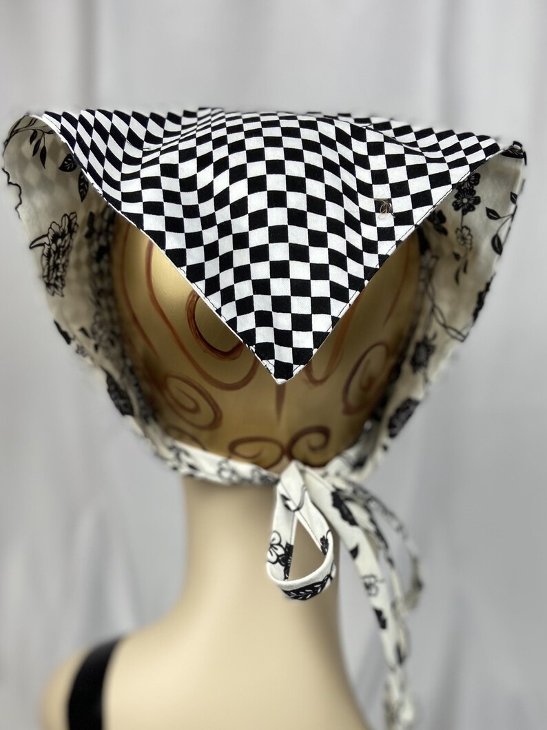 May include: A black and white checkered bonnet with a floral patterned lining. The bonnet is tied with matching fabric ties. A small metal emblem is visible on the front. The bonnet is displayed on a mannequin head.