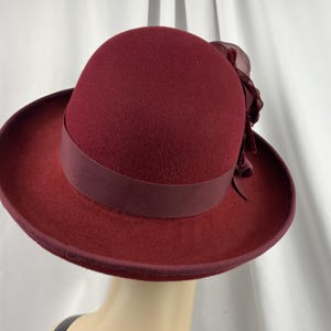 May include: A deep burgundy felt hat featuring a rounded crown and a structured brim. A matching grosgrain ribbon band encircles the base of the crown, accented on the side by a large, layered fabric flower in the same rich, dark red tone.