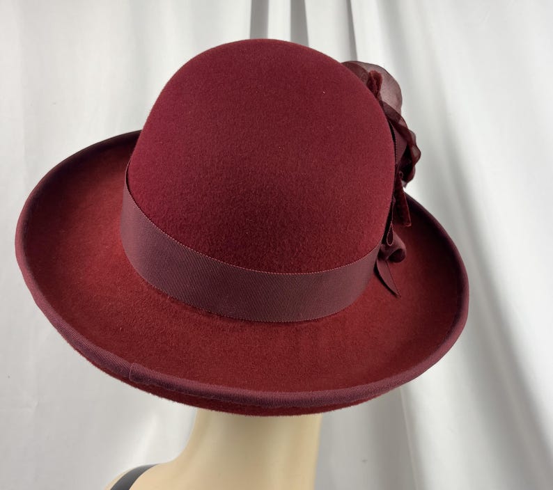 May include: A deep burgundy felt hat featuring a rounded crown and a structured brim. A matching grosgrain ribbon band encircles the base of the crown, accented on the side by a large, layered fabric flower in the same rich, dark red tone.