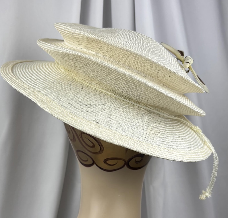 May include: A cream-colored straw hat featuring a multi-tiered, layered crown design with a wide, slightly downturned brim. The hat is adorned with a decorative side accent consisting of a small bow and a dangling, thin cord detail on the right side.
