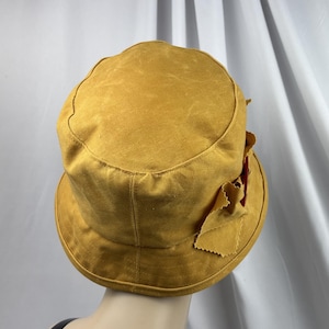Puede incluir: Un sombrero de pescador color amarillo mostaza con una parte superior plana y un ala inclinada hacia abajo. El sombrero presenta una superficie de tela texturizada y un detalle de lazo decorativo en el lateral con bordes deshilachados.