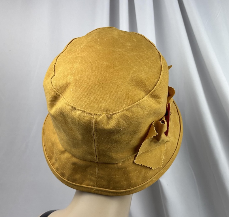 Puede incluir: Un sombrero de pescador color amarillo mostaza con una parte superior plana y un ala inclinada hacia abajo. El sombrero presenta una superficie de tela texturizada y un detalle de lazo decorativo en el lateral con bordes deshilachados.