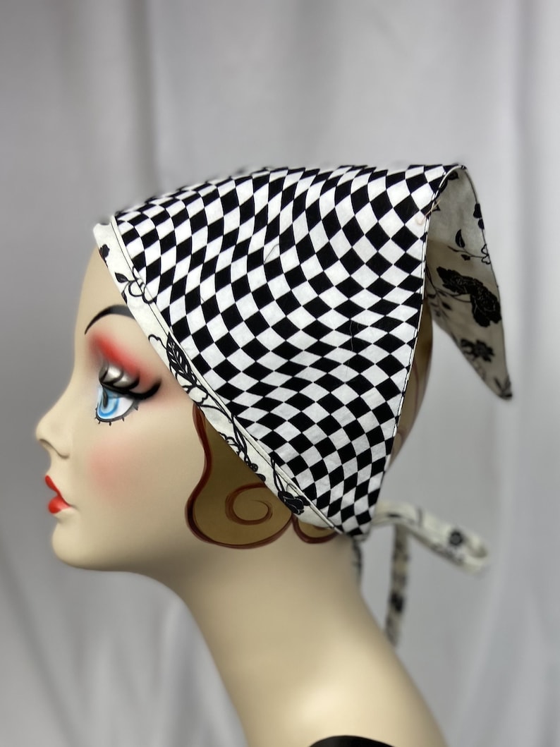 May include: A black and white checkered bandana is displayed on a mannequin head. The bandana has a classic checkerboard pattern on the front and a floral design on the edges and ties. The ties are visible on the side of the mannequin.