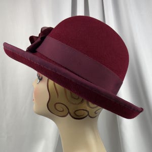 May include: A deep burgundy felt cloche hat featuring a matching wide grosgrain ribbon band and a decorative fabric flower accent on the side. The hat has a rounded crown and a slightly downturned brim, providing a structured and classic silhouette.