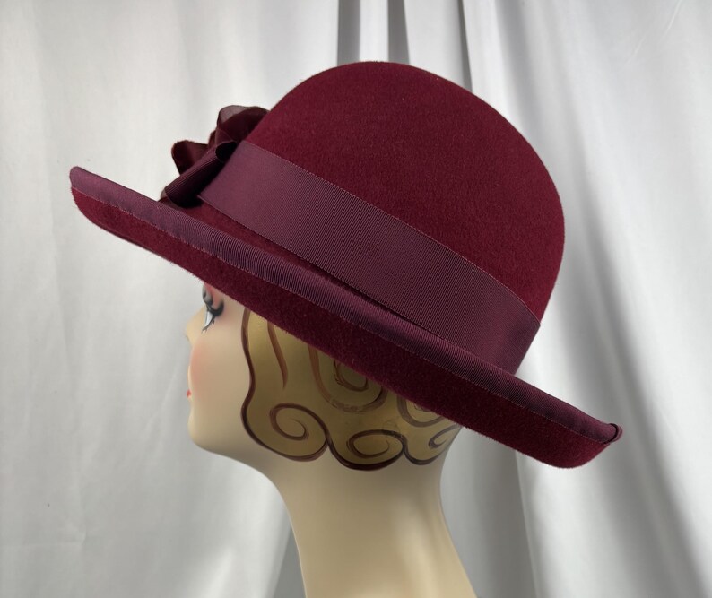 May include: A deep burgundy felt cloche hat featuring a matching wide grosgrain ribbon band and a decorative fabric flower accent on the side. The hat has a rounded crown and a slightly downturned brim, providing a structured and classic silhouette.