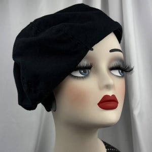 May include: A solid black beret with visible tonal stitching details along the crown, worn on a mannequin head featuring pale skin, blue eyes, long dark eyelashes, and bright red lips, presented against a plain white draped fabric background.
