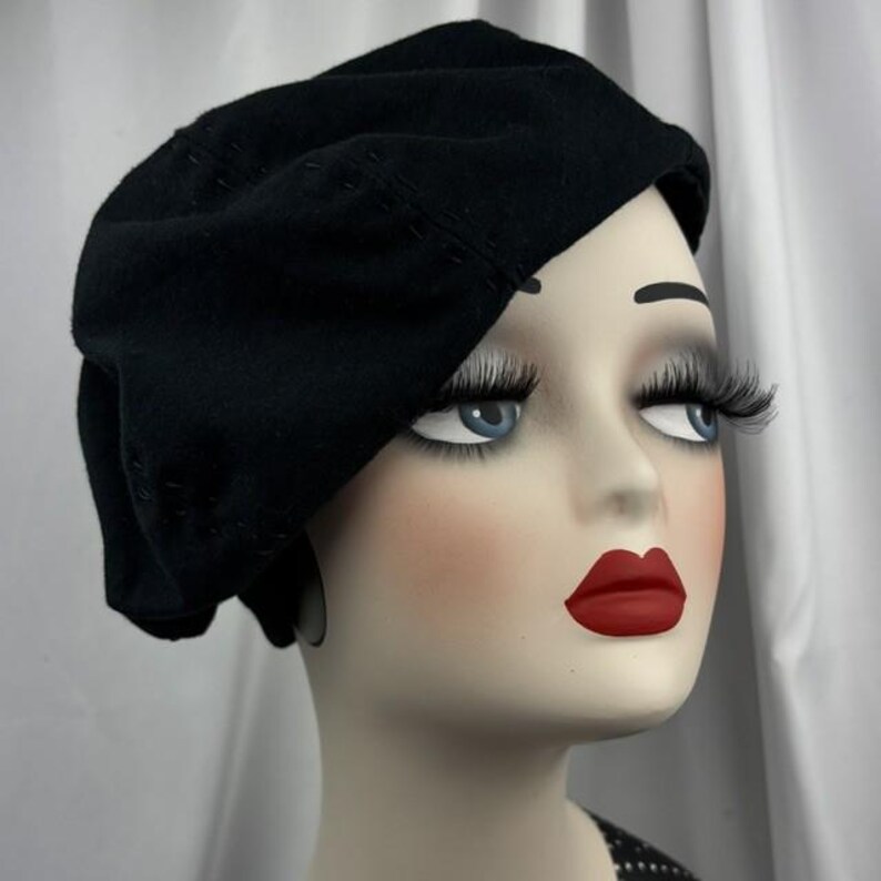 May include: A solid black beret with visible tonal stitching details along the crown, worn on a mannequin head featuring pale skin, blue eyes, long dark eyelashes, and bright red lips, presented against a plain white draped fabric background.