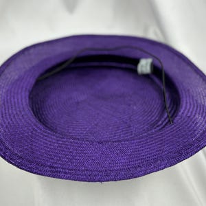 May include: A close-up of the inside of a purple sun hat. The hat is made of woven material and has a round crown and a wide brim. A black string is attached to the inside of the hat. The hat is a vibrant shade of purple.