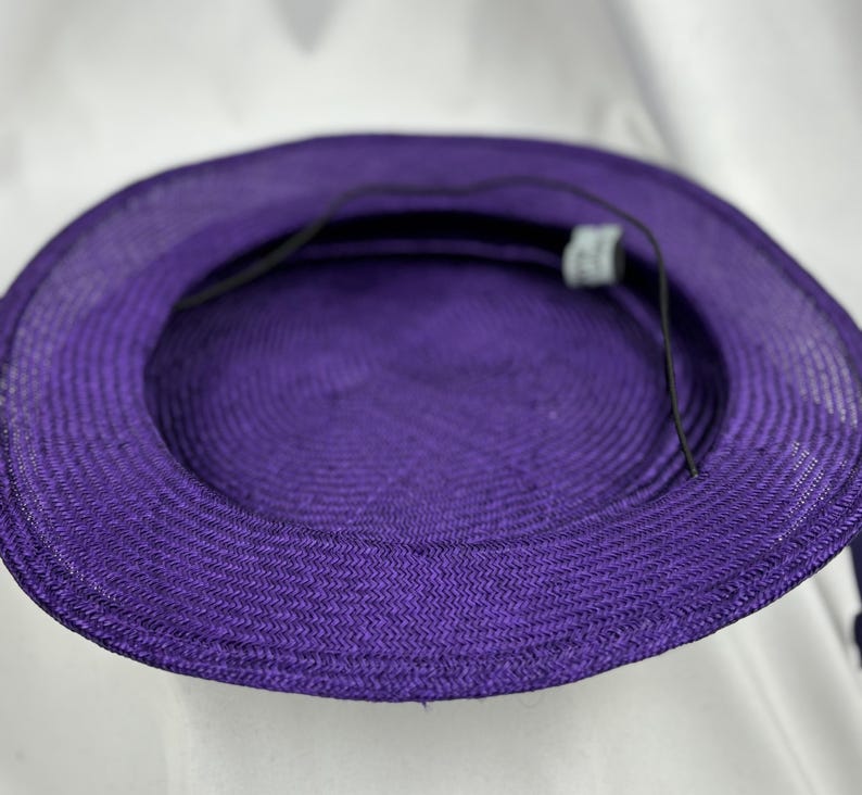 May include: A close-up of the inside of a purple sun hat. The hat is made of woven material and has a round crown and a wide brim. A black string is attached to the inside of the hat. The hat is a vibrant shade of purple.