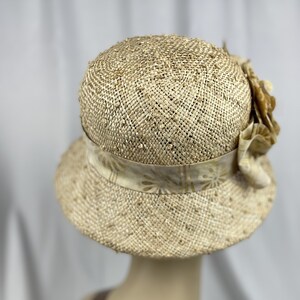 Puede incluir: Un sombrero de paja de ala ancha con una cinta de color crema con un patr&oacute;n floral. El sombrero tiene un adorno de flor marr&oacute;n en el lateral.
